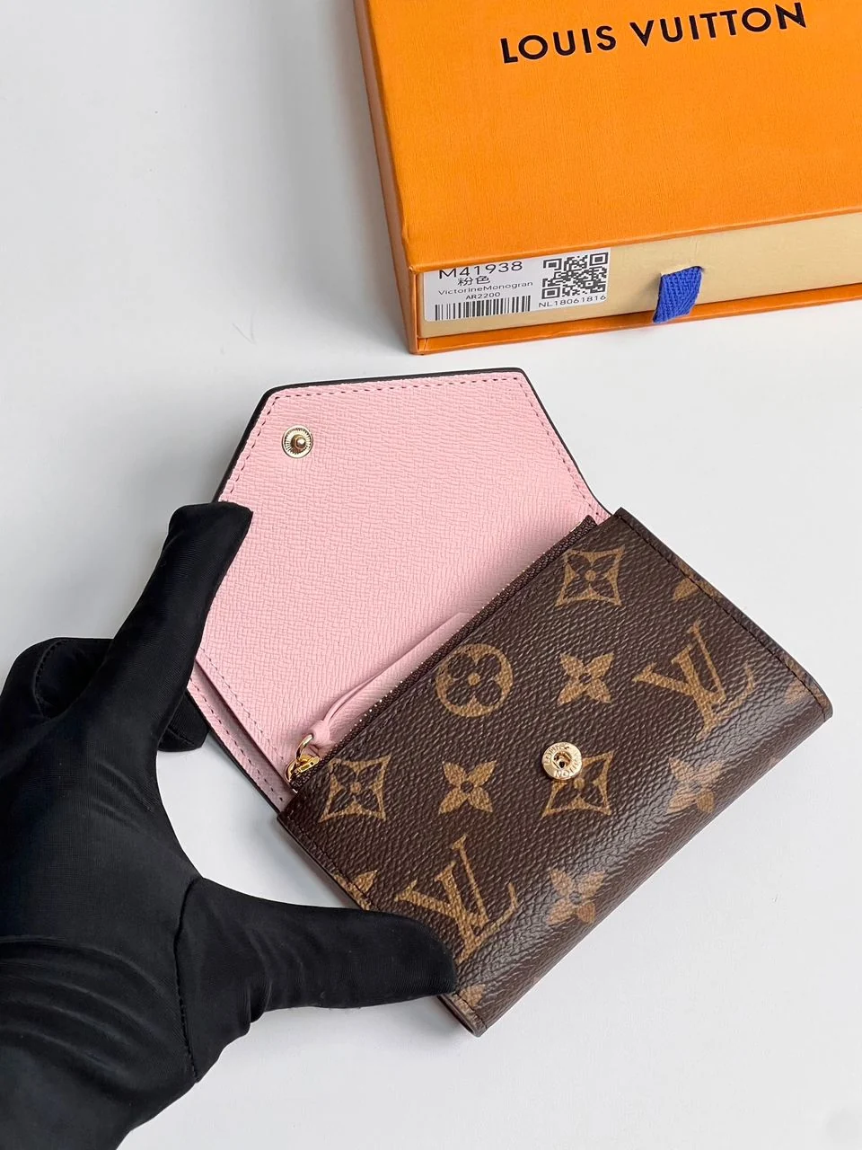 Carteira marrom monogramada com interior rosa, segurada por uma mão de luva preta diante de caixa laranja.