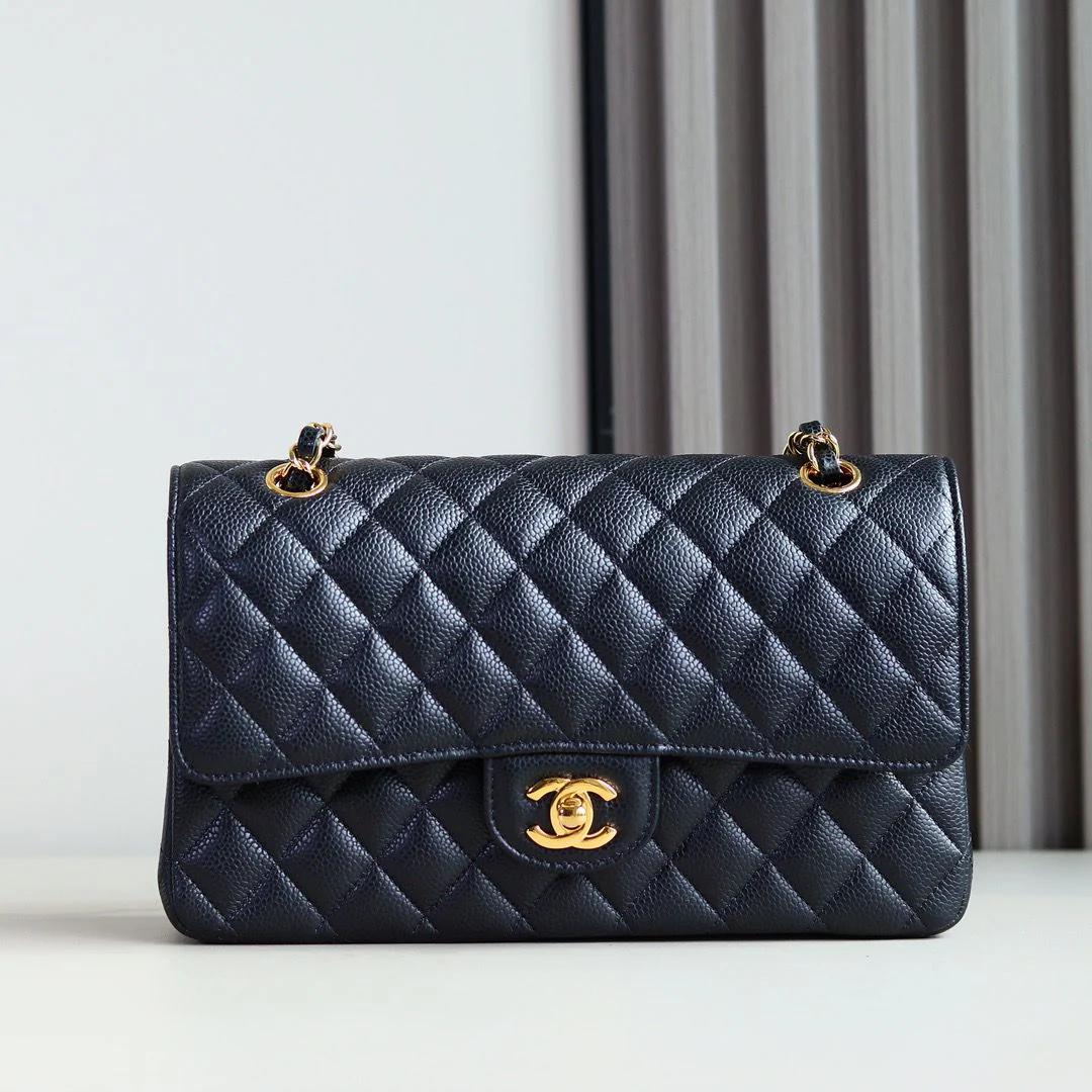 Bolsa preta matelassê com corrente e fecho dourado, sobre superfície clara e fundo de cortina.