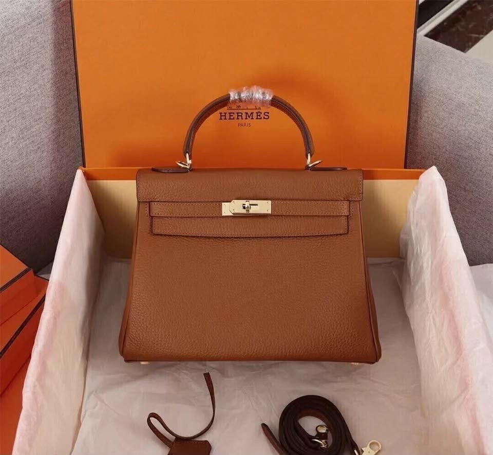Bolsa caramelo estruturada com ferragens douradas, dentro da caixa laranja aberta.