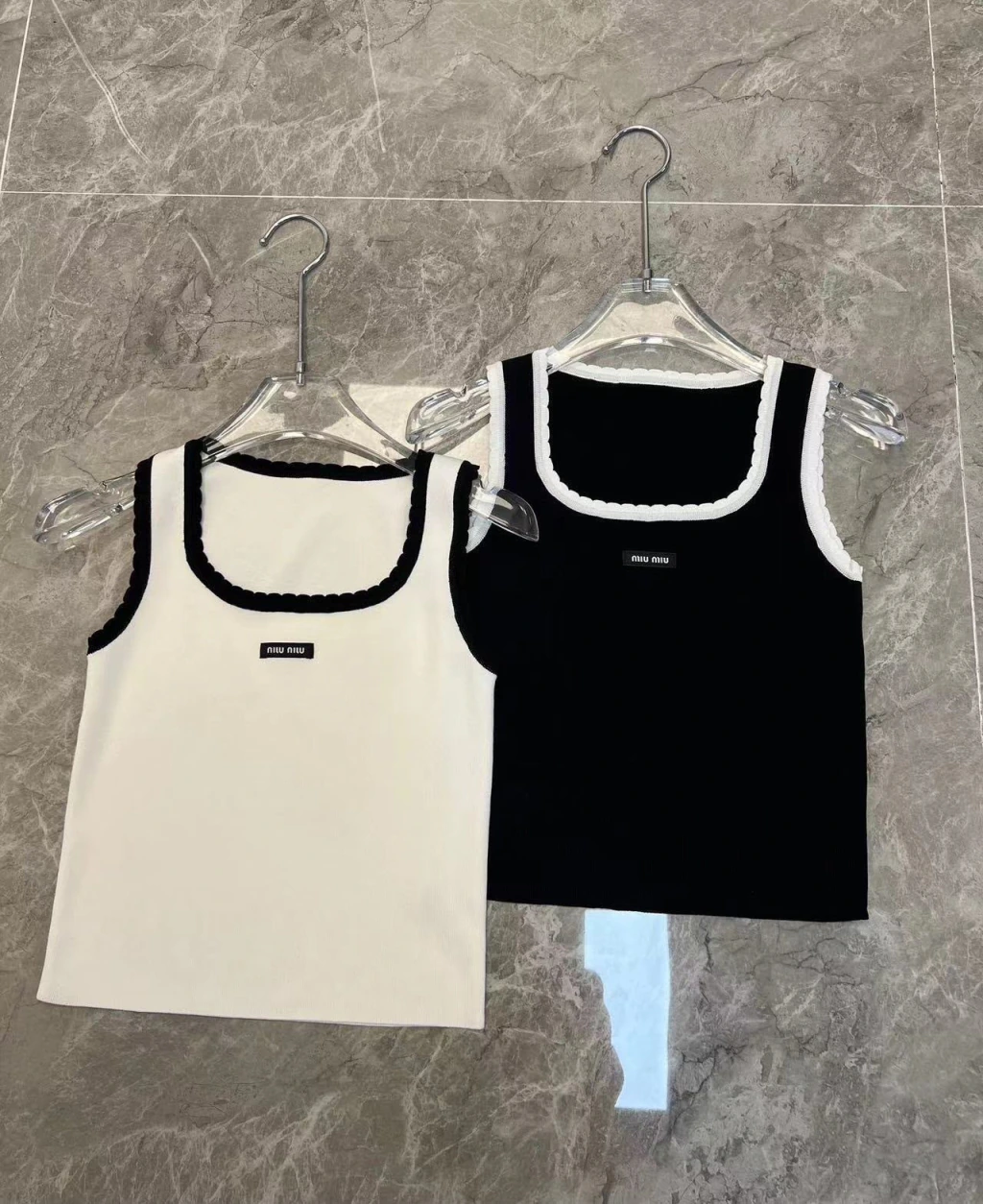 Duas regatas de malha, branca e preta com contornos contrastantes, em cabides transparentes sobre mármore.