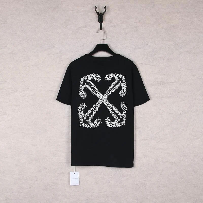 Camiseta preta vista de costas, com grande estampa branca de setas cruzadas, pendurada em cabide diante de parede clara.